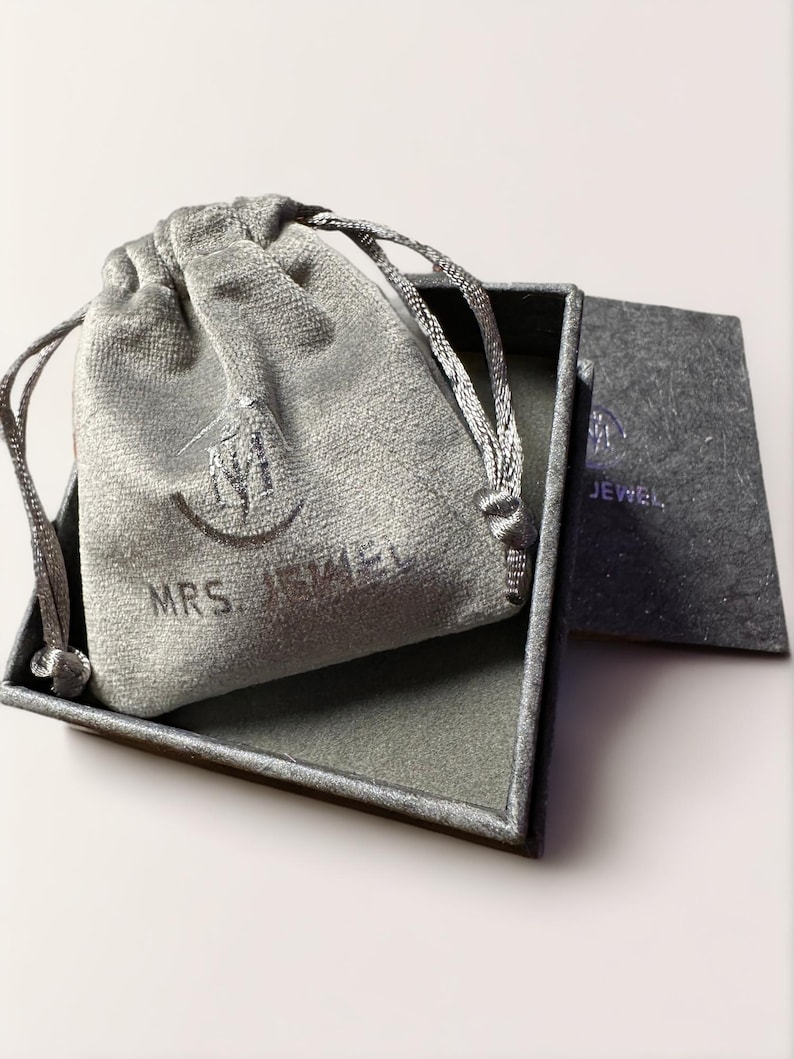 May include: A gray velvet jewelry pouch with the text "MRS. JEWEL" on it, inside a gray square box with a gray velvet lining. The box has a gray paper insert with the text "MRS. JEWEL" on it.