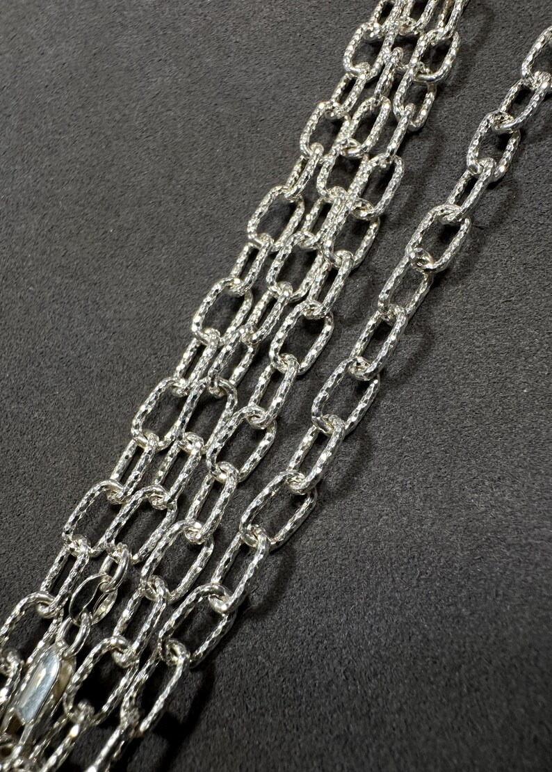 Pode incluir: Um colar de corrente de prata com um padr&atilde;o texturizado, semelhante a um diamante. A corrente &eacute; feita de v&aacute;rias fitas de elos ovais interligados.