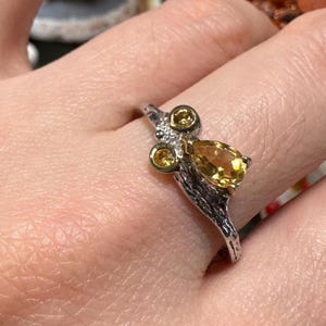 May include: A silver ring with a nature-inspired design. The ring features three small, round yellow gemstones and a larger, pear-shaped yellow gemstone. The band has a textured, branch-like appearance.
