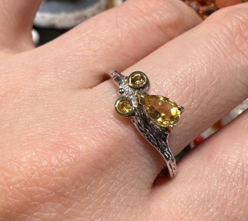 May include: A silver ring with a nature-inspired design. The ring features three small, round yellow gemstones and a larger, pear-shaped yellow gemstone. The band has a textured, branch-like appearance.