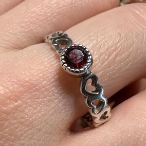 May include: A silver ring with a central red gemstone and a band featuring heart-shaped cutouts. The gemstone is round and set in a beaded bezel. The ring is worn on a finger.