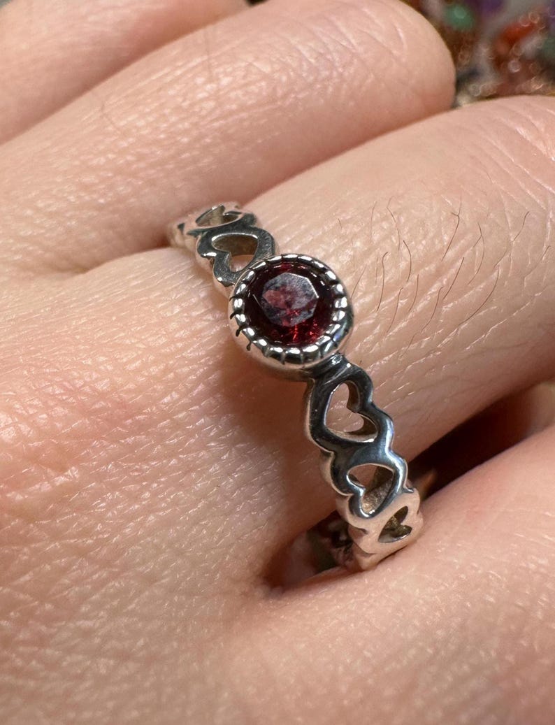 May include: A silver ring with a central red gemstone and a band featuring heart-shaped cutouts. The gemstone is round and set in a beaded bezel. The ring is worn on a finger.