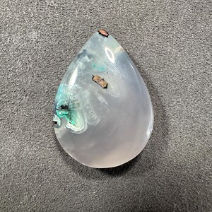 Puede incluir: Un cabuj&oacute;n de &aacute;gata rosa en forma de l&aacute;grima con inclusiones verdes y marrones. La piedra es transl&uacute;cida y tiene una superficie lisa y pulida.