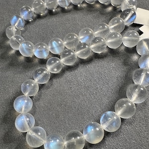 Könnte beinhalten: Eine Kette oder Armband aus klaren, runden Perlen. Jede Perle hat einen Durchmesser von etwa 0,8 cm. Die Perlen sind von transluzenter weißer Farbe mit schimmernden blauen Reflexen. Die Perlen sind aufgereiht und bilden eine Schleife.