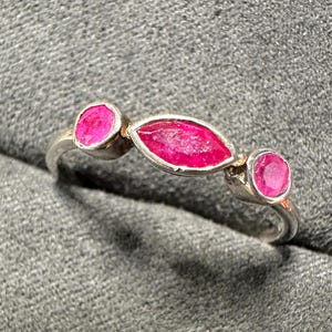 May include: A silver ring featuring three vibrant, oval-cut ruby gemstones. The central ruby is elongated, flanked by two round rubies, all set in a polished silver band. The ring's design is simple yet elegant, showcasing the rich color of the gemstones.