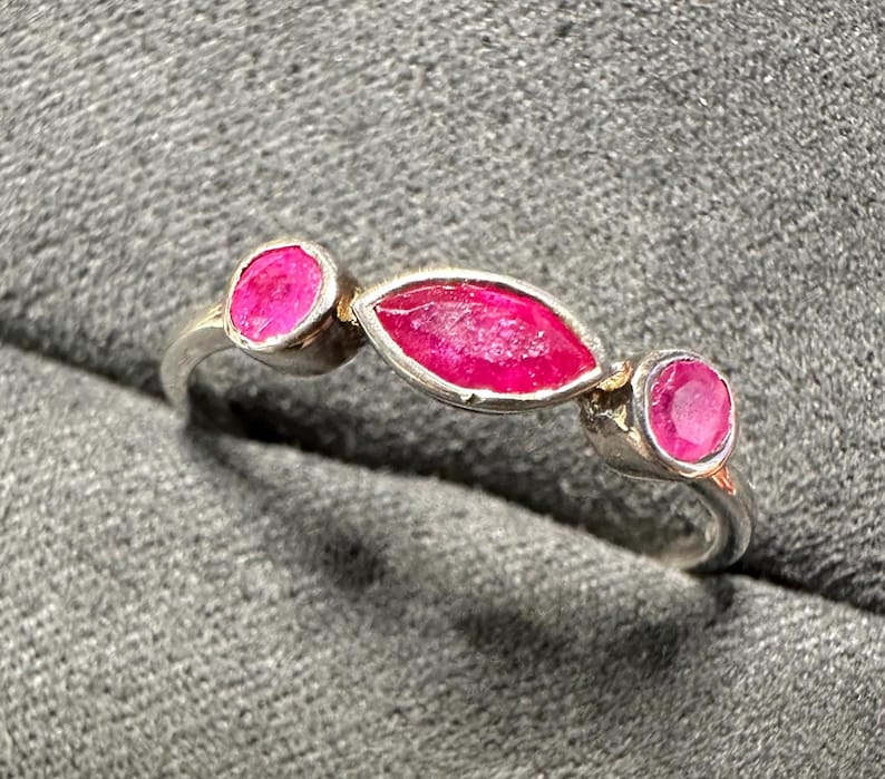 May include: A silver ring featuring three vibrant, oval-cut ruby gemstones. The central ruby is elongated, flanked by two round rubies, all set in a polished silver band. The ring's design is simple yet elegant, showcasing the rich color of the gemstones.