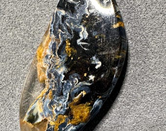 May include: A polished, teardrop-shaped gemstone with a striking pattern. The stone features swirling veins of blue, gold, and white against a dark background. The gemstone is set against a gray surface.