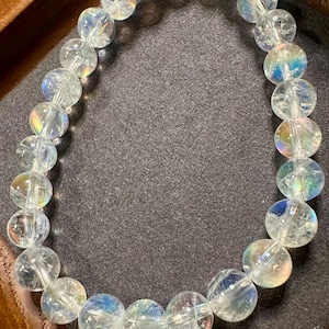 Pode incluir: Uma pulseira de contas de quartzo transparente com flashes iridescentes de cor. As contas redondas s&atilde;o enfiadas juntas, criando uma forma circular. A pulseira &eacute; exibida em uma superf&iacute;cie cinza escuro, dentro de uma bandeja de madeira.