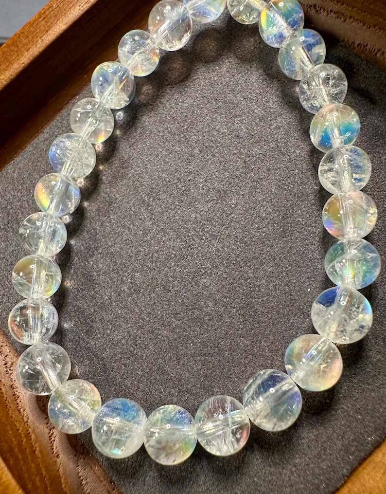 Pode incluir: Uma pulseira de contas de quartzo transparente com flashes iridescentes de cor. As contas redondas s&atilde;o enfiadas juntas, criando uma forma circular. A pulseira &eacute; exibida em uma superf&iacute;cie cinza escuro, dentro de uma bandeja de madeira.