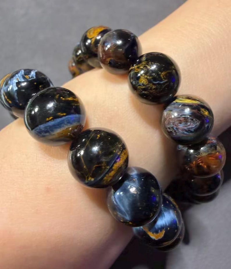 May include: A bracelet featuring large, round beads with a polished, glossy finish. The beads display a mix of dark blue, black, and golden-brown colors, creating a marbled effect. The bracelet is strung together, showcasing the natural stone's unique patterns.