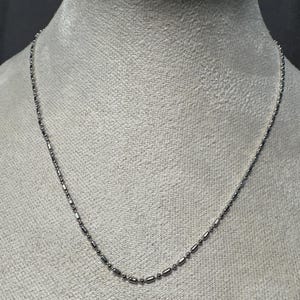 May include: A delicate silver necklace with a repeating pattern of small, elongated beads and round beads. The necklace is displayed on a mannequin, showcasing its length and design. The chain is thin and appears to be made of metal.