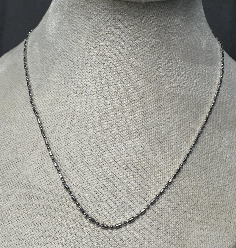 May include: A delicate silver necklace with a repeating pattern of small, elongated beads and round beads. The necklace is displayed on a mannequin, showcasing its length and design. The chain is thin and appears to be made of metal.