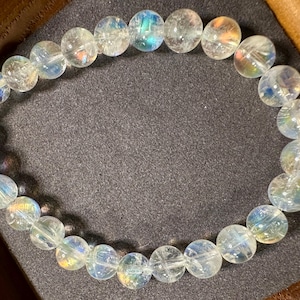 Pode incluir: Uma pulseira de contas claras e iridescentes. As contas redondas exibem um sutil efeito arco-&iacute;ris, com tons de azul, amarelo e verde. A pulseira &eacute; apresentada em uma superf&iacute;cie cinza escuro, destacando a qualidade luminosa das contas.