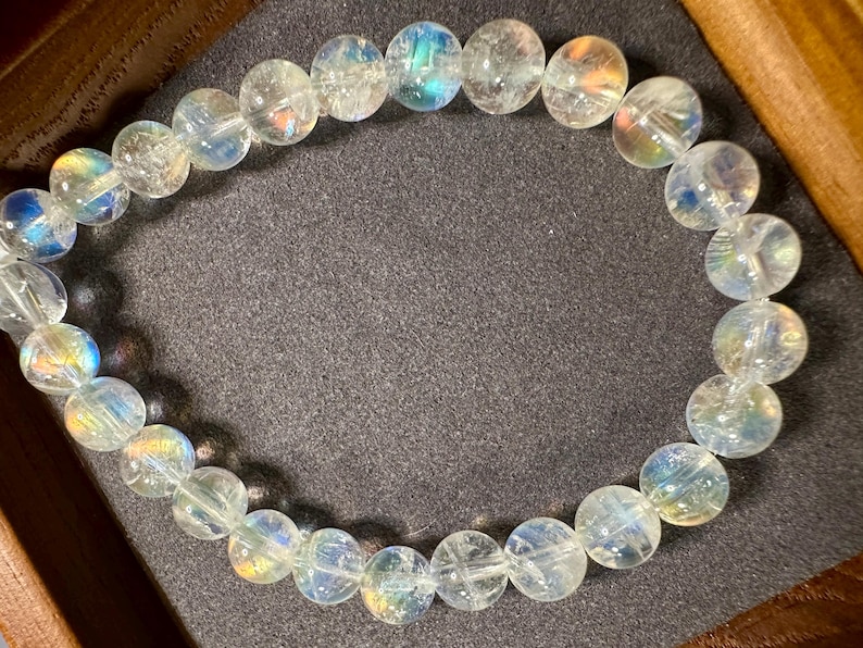 Pode incluir: Uma pulseira de contas claras e iridescentes. As contas redondas exibem um sutil efeito arco-&iacute;ris, com tons de azul, amarelo e verde. A pulseira &eacute; apresentada em uma superf&iacute;cie cinza escuro, destacando a qualidade luminosa das contas.