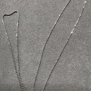 May include: A delicate silver necklace with a ball chain design. The necklace is displayed on a gray surface. The chain is thin and features small, evenly spaced beads. The clasp is visible at the top.