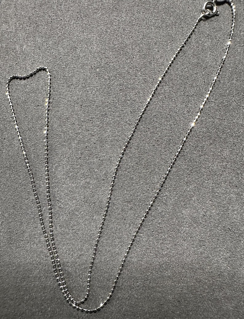 May include: A delicate silver necklace with a ball chain design. The necklace is displayed on a gray surface. The chain is thin and features small, evenly spaced beads. The clasp is visible at the top.