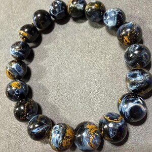 May include: A beaded bracelet featuring round, polished beads. The beads are primarily dark blue with swirling patterns of lighter blue and gold. The bracelet is displayed on a gray surface, showcasing the natural colors and textures of the gemstones.
