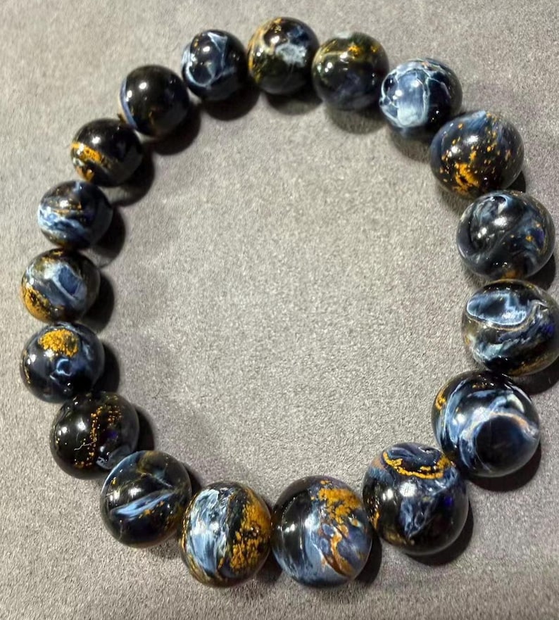 May include: A beaded bracelet featuring round, polished beads. The beads are primarily dark blue with swirling patterns of lighter blue and gold. The bracelet is displayed on a gray surface, showcasing the natural colors and textures of the gemstones.
