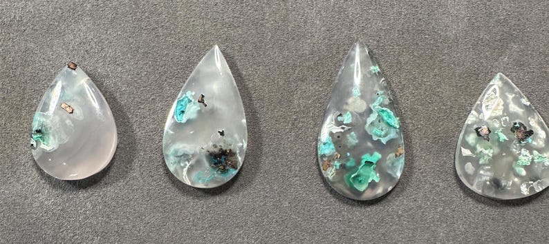Puede incluir: Cuatro cabujones en forma de l&aacute;grima de cuarzo transparente con inclusiones de minerales verdes y marrones. Los cabujones est&aacute;n pulidos y tienen un acabado liso y brillante.