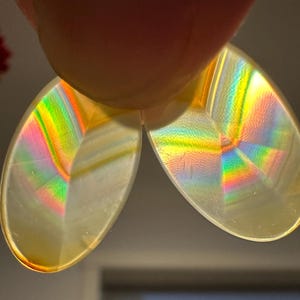 May include: Two iridescent, teardrop-shaped objects are held up, reflecting a spectrum of rainbow colors. The objects appear to be made of a translucent material, with a pale yellow hue. The light refracts, creating a vibrant display of colors.