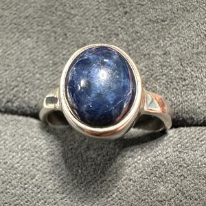 May include: A silver ring featuring a large, oval-shaped, deep blue gemstone. The gemstone is set in a silver bezel and the ring has a simple, elegant design. The ring is a classic piece of jewelry.