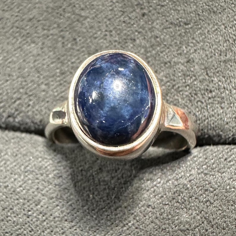 May include: A silver ring featuring a large, oval-shaped, deep blue gemstone. The gemstone is set in a silver bezel and the ring has a simple, elegant design. The ring is a classic piece of jewelry.