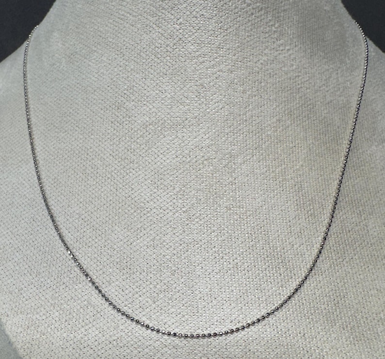 May include: A delicate silver-colored ball chain necklace. The necklace is displayed against a neutral background, highlighting its simple and elegant design. This jewelry piece is suitable for everyday wear.