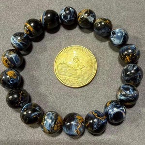 May include: A beaded bracelet featuring dark blue and gold-colored Pietersite gemstones. The bracelet is circular, with each bead displaying unique patterns. A Canadian one-dollar coin is placed in the center for size comparison.