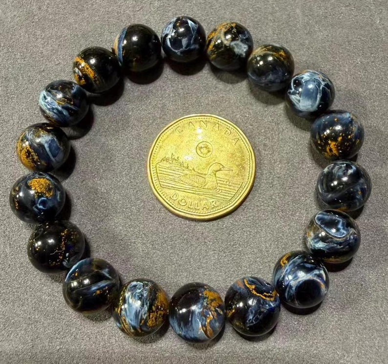 May include: A beaded bracelet featuring dark blue and gold-colored Pietersite gemstones. The bracelet is circular, with each bead displaying unique patterns. A Canadian one-dollar coin is placed in the center for size comparison.