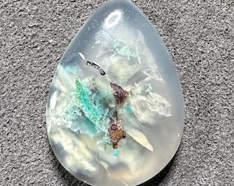 May include: A teardrop-shaped cabochon of white agate with inclusions of green, blue, and brown minerals. The stone is translucent and has a smooth, polished surface.