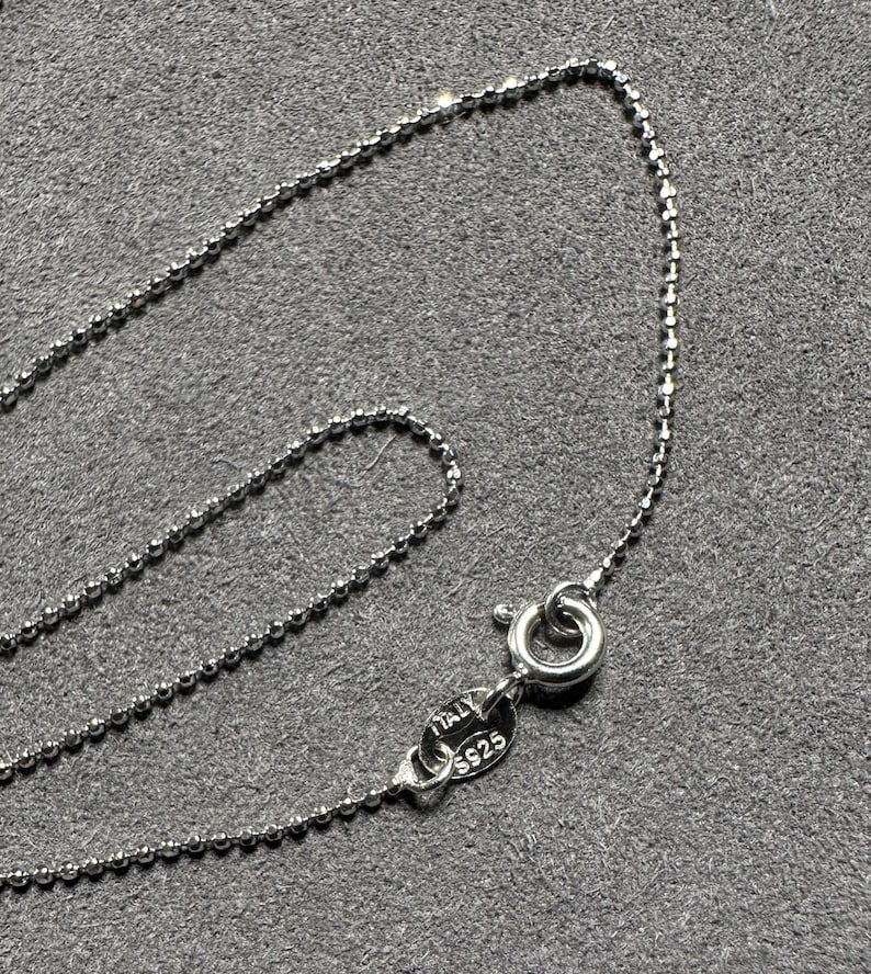 May include: A delicate silver ball chain necklace with a lobster clasp. The clasp is stamped with "ITALY S925". The necklace is displayed on a gray surface.