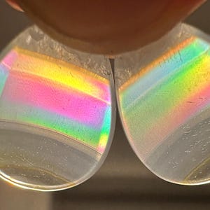 May include: Two clear, teardrop-shaped objects refracting light, displaying a rainbow spectrum of colours. The colours shift from pink to yellow to green. The objects are held by a person's fingers.