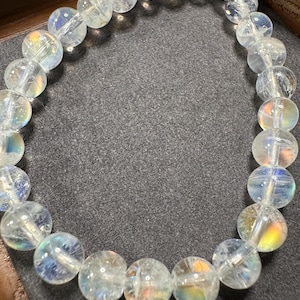 Pode incluir: Uma pulseira de contas clara e iridescente. As contas redondas t&ecirc;m uma qualidade transl&uacute;cida, com sutis flashes de cores de arco-&iacute;ris. A pulseira &eacute; amarrada, criando uma forma circular flex&iacute;vel. As contas t&ecirc;m aproximadamente 1,3 cm de di&acirc;metro.