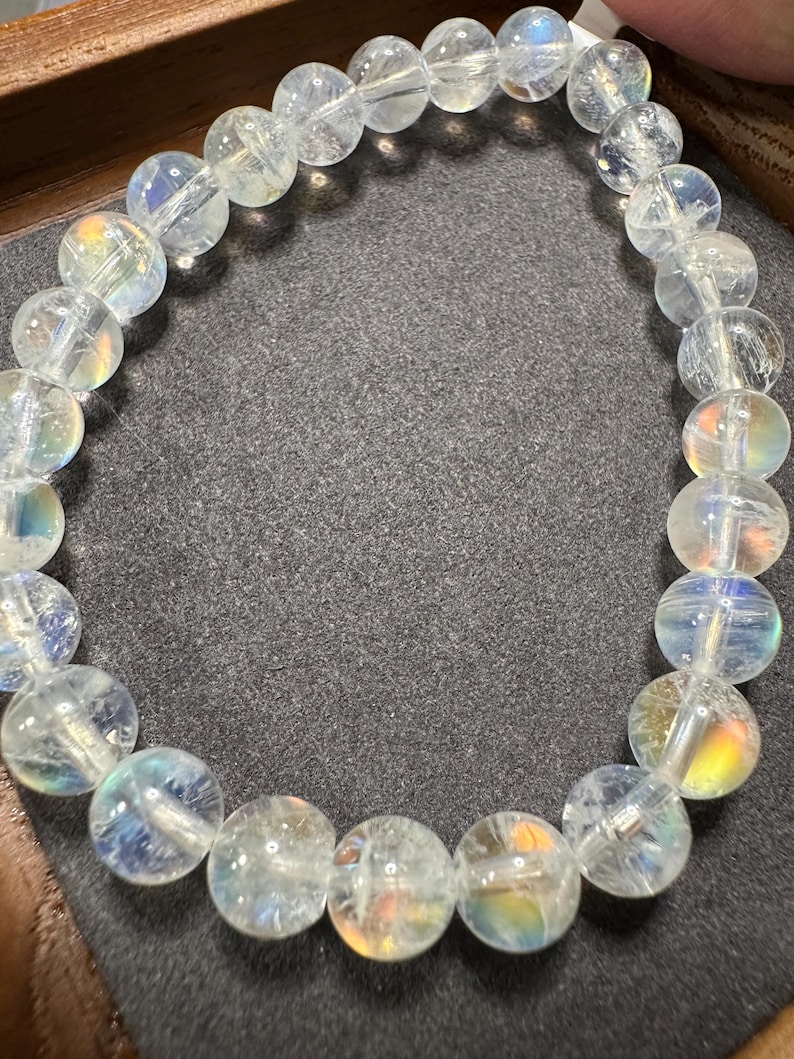 Pode incluir: Uma pulseira de contas clara e iridescente. As contas redondas t&ecirc;m uma qualidade transl&uacute;cida, com sutis flashes de cores de arco-&iacute;ris. A pulseira &eacute; amarrada, criando uma forma circular flex&iacute;vel. As contas t&ecirc;m aproximadamente 1,3 cm de di&acirc;metro.