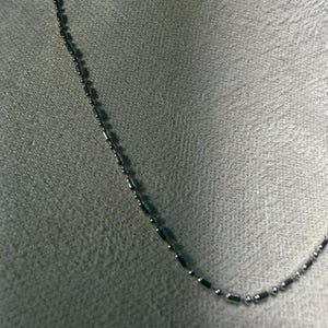 May include: A delicate silver-colored necklace with a chain of small, cylindrical beads and round beads. The necklace is displayed against a neutral gray background, highlighting its simple elegance. The chain is approximately 18 inches long.