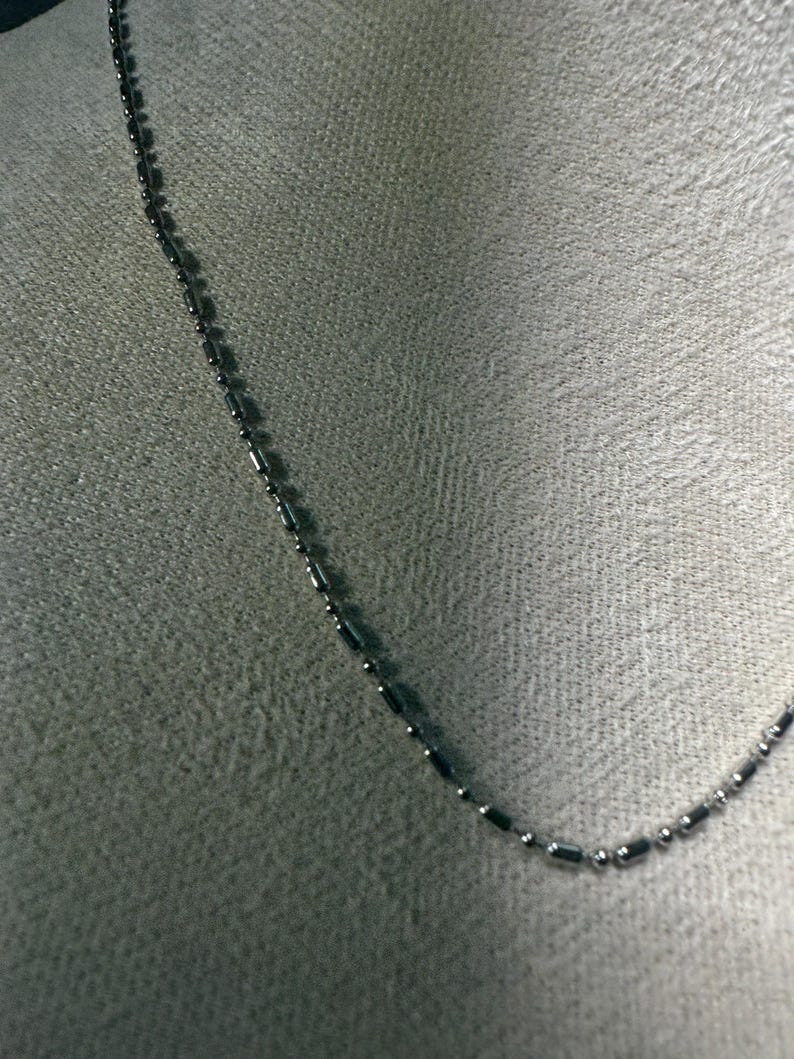 May include: A delicate silver-colored necklace with a chain of small, cylindrical beads and round beads. The necklace is displayed against a neutral gray background, highlighting its simple elegance. The chain is approximately 18 inches long.
