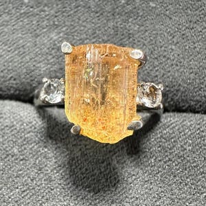 May include: A silver ring featuring a large, rectangular, amber-colored gemstone as the centerpiece, flanked by two smaller, clear gemstones. The ring is set against a textured gray background, highlighting the ring's design and the gemstones' brilliance.