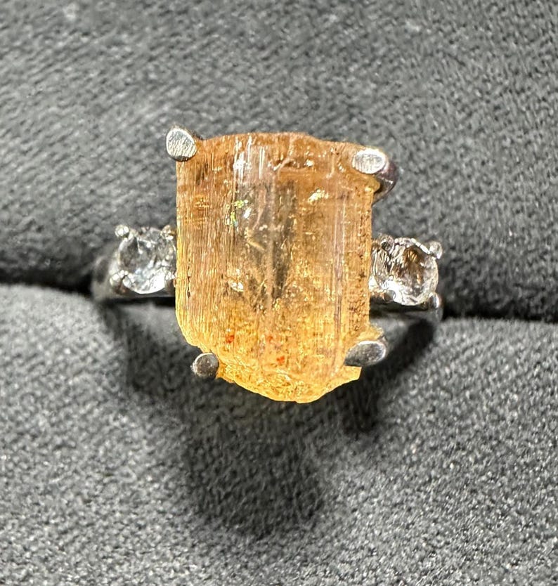 May include: A silver ring featuring a large, rectangular, amber-colored gemstone as the centerpiece, flanked by two smaller, clear gemstones. The ring is set against a textured gray background, highlighting the ring's design and the gemstones' brilliance.
