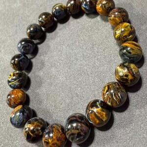 May include: A beaded bracelet featuring polished, round tiger eye stones. The stones display a mix of golden brown, dark brown, and blue hues. The bracelet is strung together, creating a circular shape.