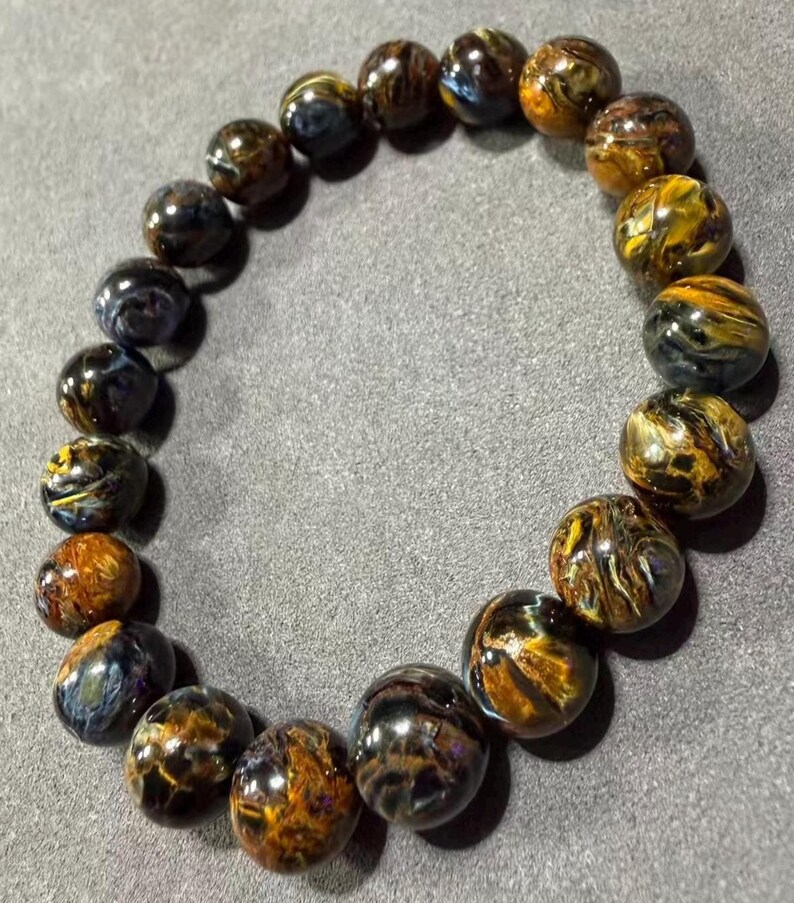 May include: A beaded bracelet featuring polished, round tiger eye stones. The stones display a mix of golden brown, dark brown, and blue hues. The bracelet is strung together, creating a circular shape.