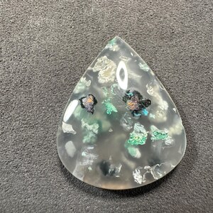 Puede incluir: Un cabuj&oacute;n de &aacute;gata transparente en forma de l&aacute;grima con inclusiones verdes, negras y plateadas. La piedra es transl&uacute;cida y tiene un acabado pulido.