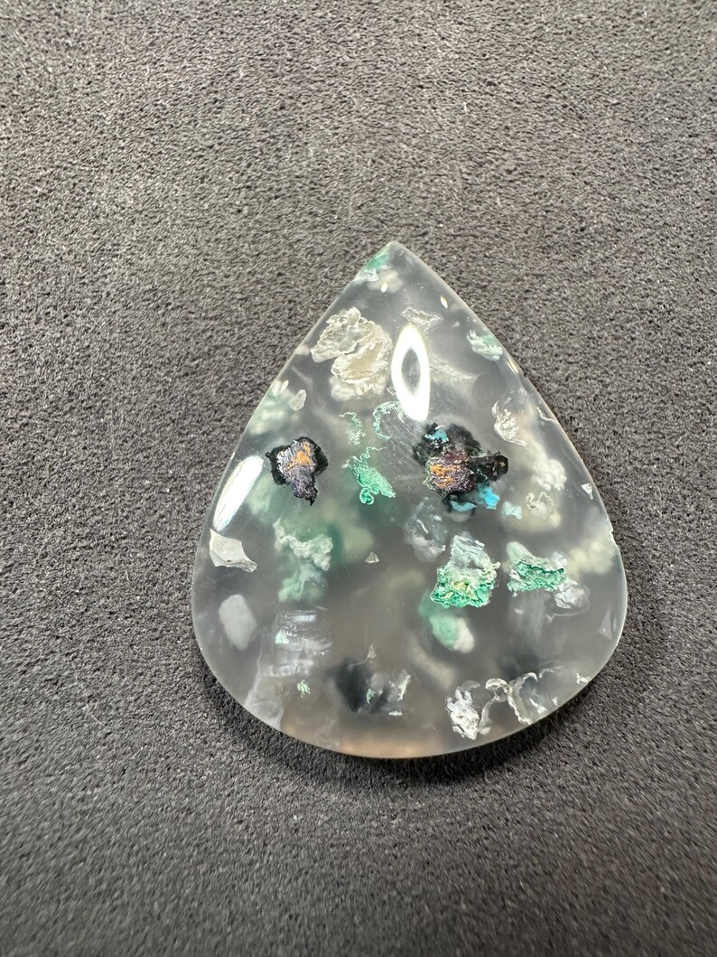 Puede incluir: Un cabuj&oacute;n de &aacute;gata transparente en forma de l&aacute;grima con inclusiones verdes, negras y plateadas. La piedra es transl&uacute;cida y tiene un acabado pulido.