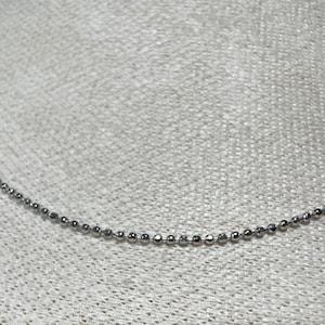 May include: A delicate silver ball chain necklace. The necklace is composed of small, evenly spaced, spherical beads. The chain is displayed against a textured, light-colored background.