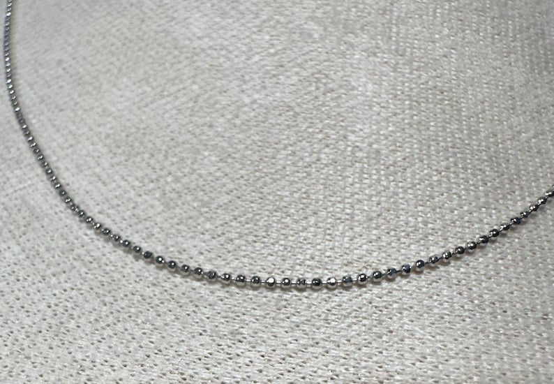 May include: A delicate silver ball chain necklace. The necklace is composed of small, evenly spaced, spherical beads. The chain is displayed against a textured, light-colored background.
