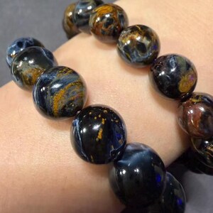 May include: A close-up of a bracelet made of polished, round beads. The beads are primarily dark blue with swirling patterns of gold and brown. The bracelet is worn on a wrist.