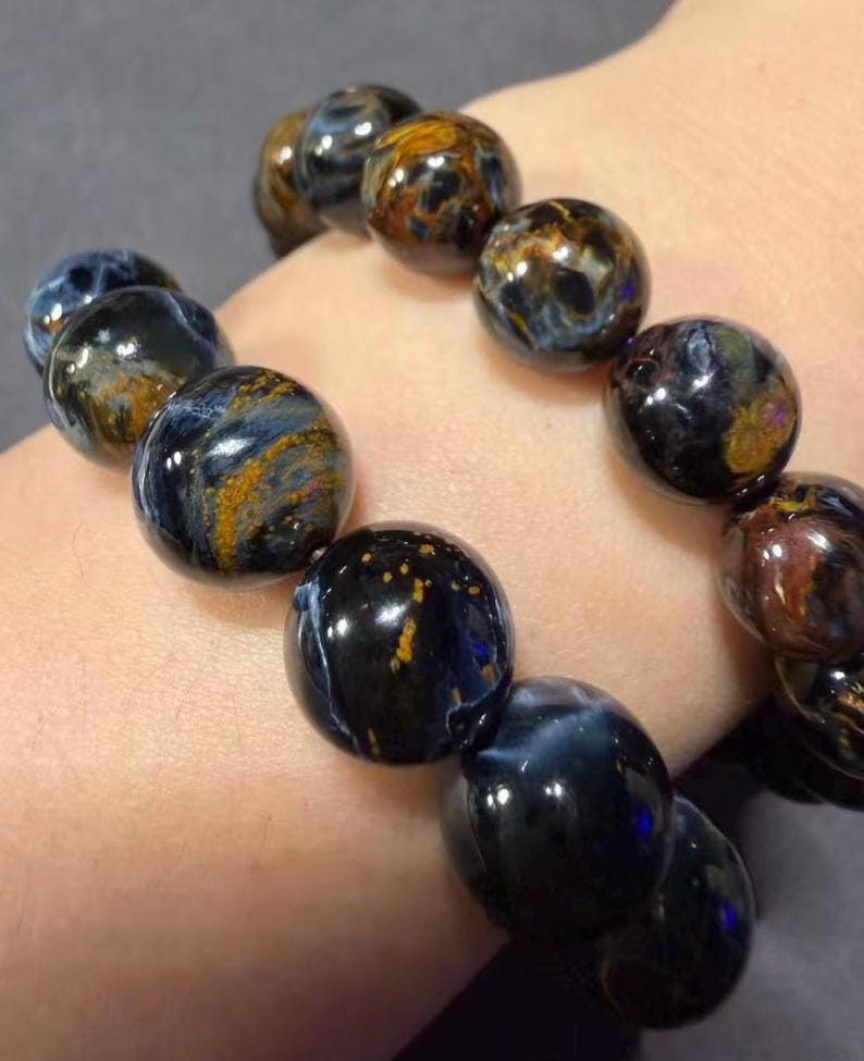 May include: A close-up of a bracelet made of polished, round beads. The beads are primarily dark blue with swirling patterns of gold and brown. The bracelet is worn on a wrist.