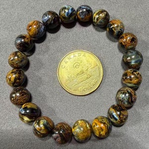 May include: A bracelet made of polished, round tiger eye beads in shades of brown, gold, and blue. A Canadian dollar coin is in the center of the bracelet. The beads are approximately 0.4 inches in diameter.