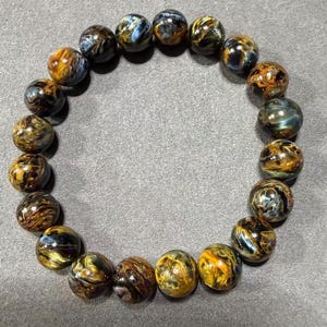 May include: A bracelet made of polished tiger's eye gemstone beads. The beads are round, displaying a mix of golden brown, blue, and black colors. The bracelet is displayed on a gray surface.