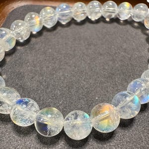 Pode incluir: Uma pulseira de contas de quartzo transparente com flashes iridescentes de arco-&iacute;ris. As contas redondas s&atilde;o enfiadas juntas, criando uma forma circular. A pulseira &eacute; exibida em uma superf&iacute;cie cinza escuro.