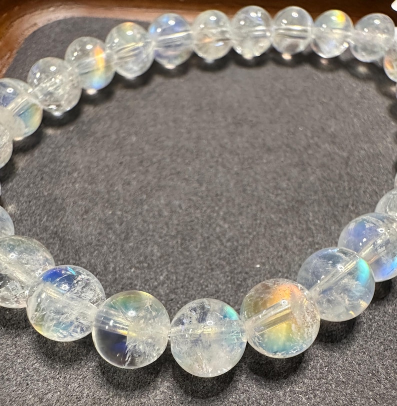 Pode incluir: Uma pulseira de contas de quartzo transparente com flashes iridescentes de arco-&iacute;ris. As contas redondas s&atilde;o enfiadas juntas, criando uma forma circular. A pulseira &eacute; exibida em uma superf&iacute;cie cinza escuro.